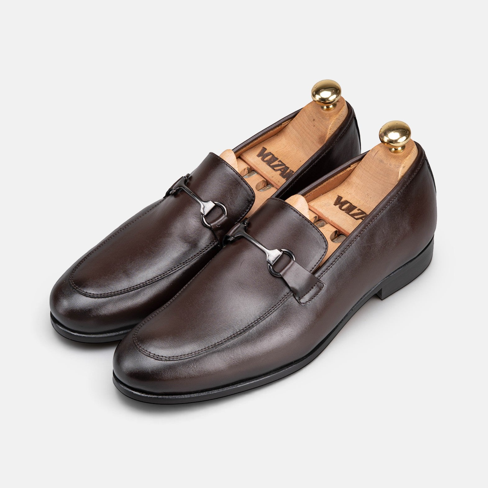 VOLZAK brown leather loafers with a metal bit detail and wooden shoe trees, presented on a light grey background.